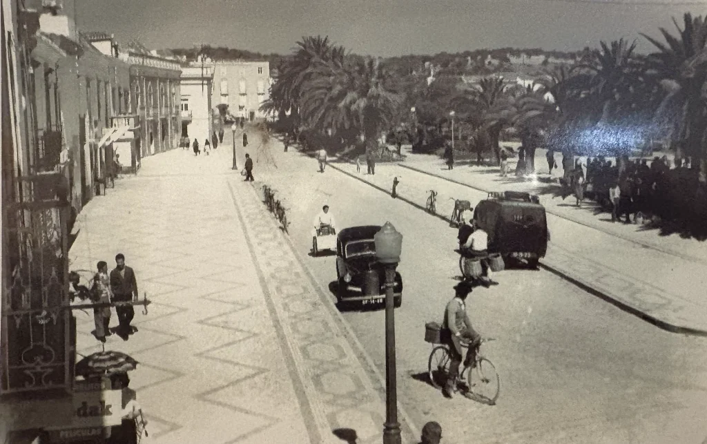Rue animée de Tavira, Portugal, années 1950, voitures anciennes et cycliste, ambiance rétro et historique.