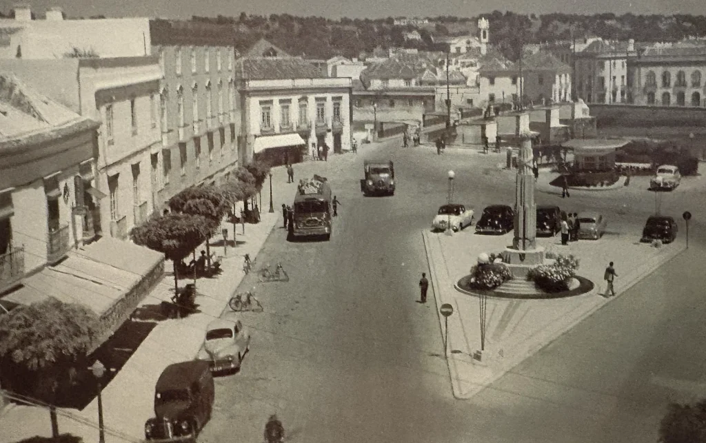 Vue ancienne de Tavira, Portugal, avec voitures d'époque et bâtiments historiques autour d'une place centrale animée.