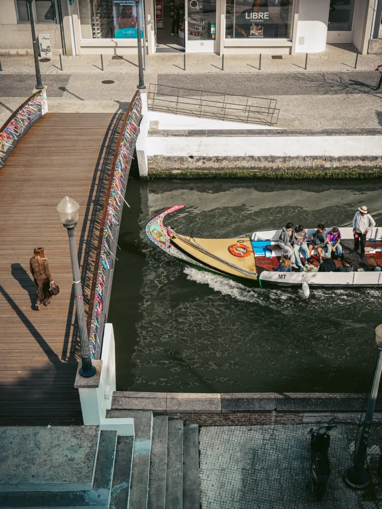 Aveiro canal with a colorful moliceiro boat and a pedestrian bridge, lively and touristy atmosphere.
