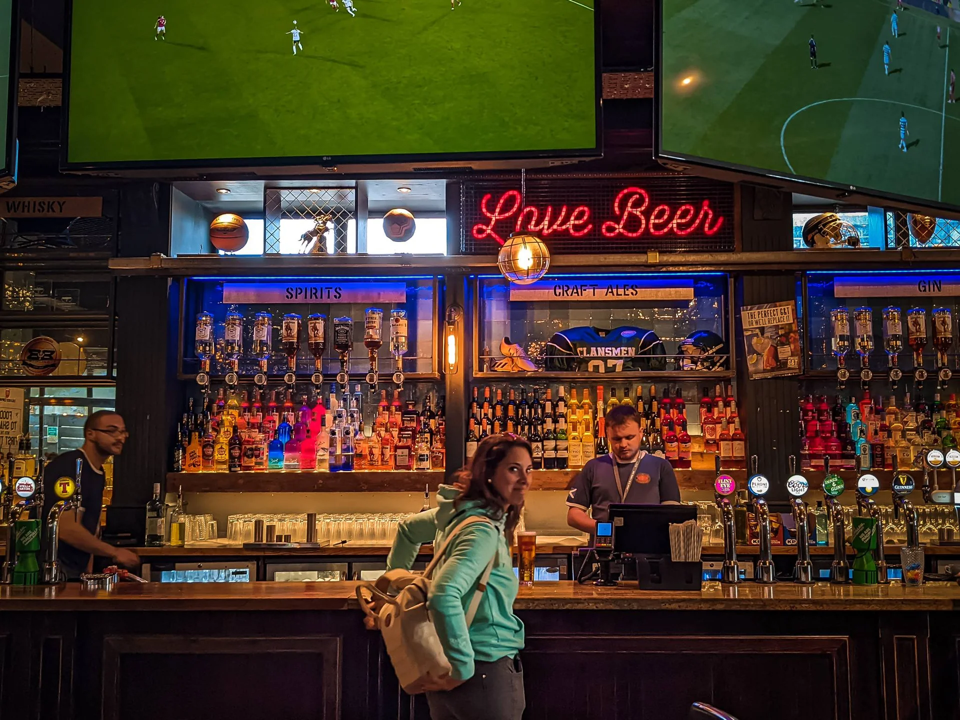 Caroline at the counter of a lively pub in Stirling with screens showing football matches