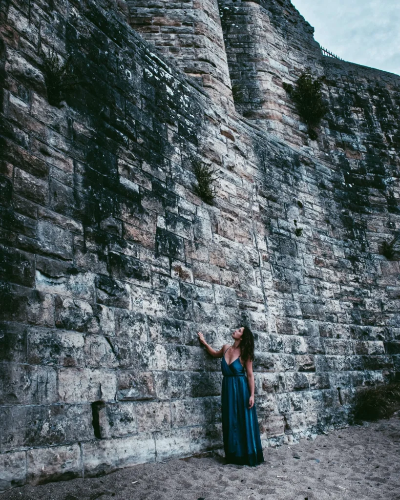 Caroline in a long blue dress in front of a large ancient stone wall in St Andrews