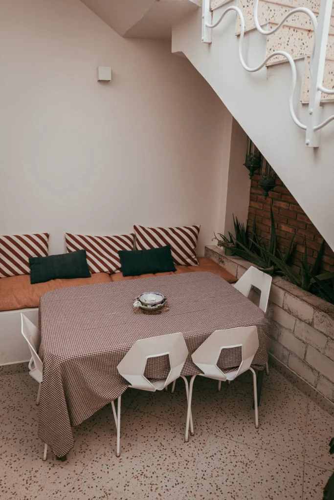 Outdoor dining area under a staircase, with a table covered with a checkered tablecloth, white chairs, and a bench with striped cushions, at Casita Erika in Fuseta, Algarve.