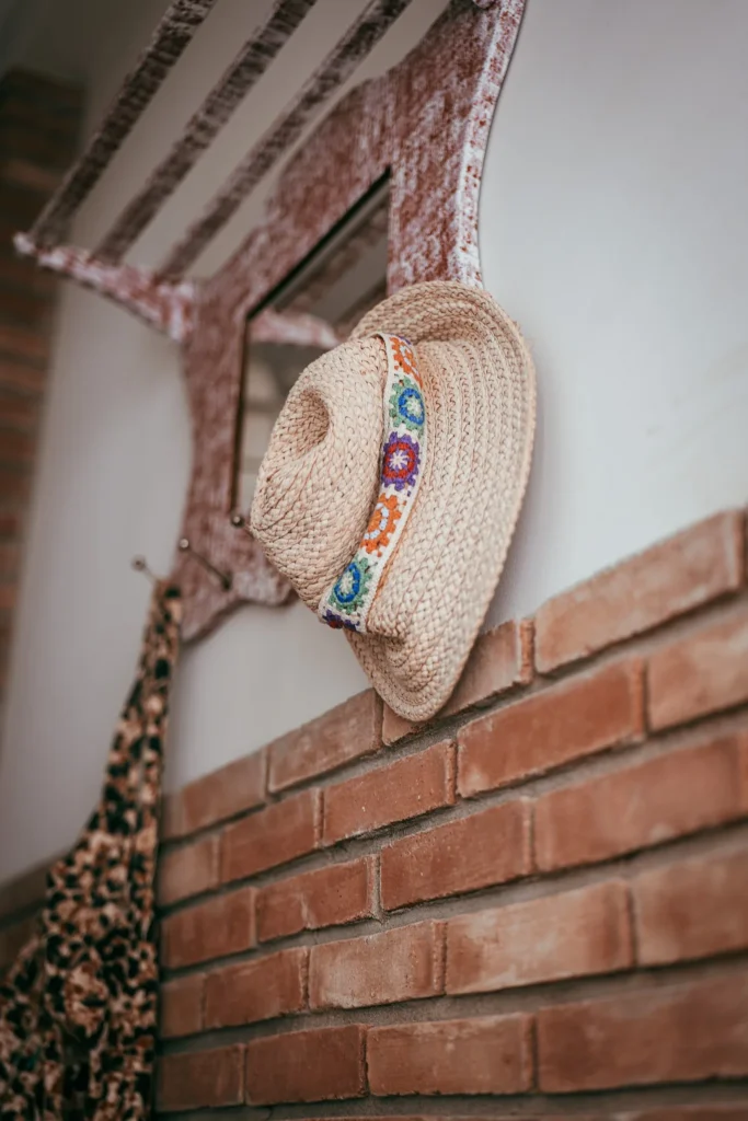 Straw hat decorated with colorful patterns hanging on a wall-mounted coat rack in an entryway with brick walls, warm interior ambiance in Fuseta, Algarve.