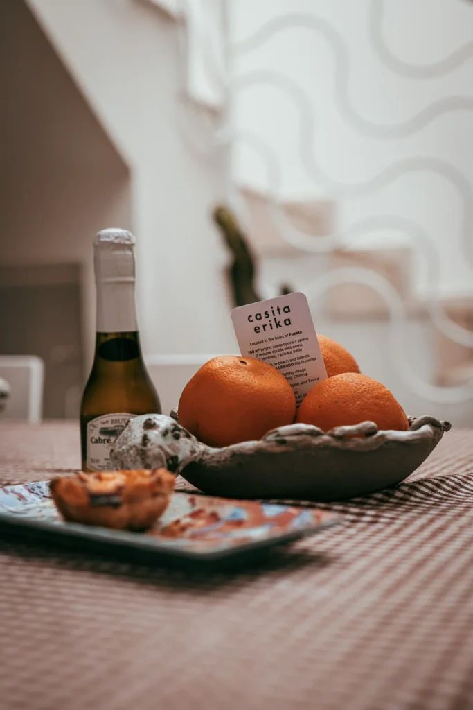 On a table in Fuseta, Algarve, a bowl of oranges, a bottle of Cabreiroa sparkling wine, and a Portuguese pastry are arranged, with a 'casita erika' card prominently displayed.