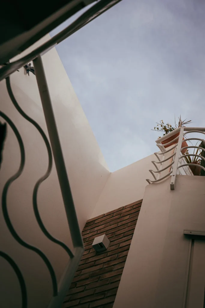 View from below of an inner courtyard with white walls and bricks, featuring a metal railing and a flower pot, under the sky of Fuseta in the Algarve.