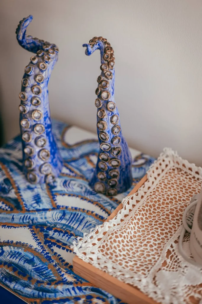 Two blue octopus tentacle-shaped sculptures are placed on a decorative fabric with marine patterns, next to a wooden tray covered with lace. Typical seaside atmosphere of the Algarve.