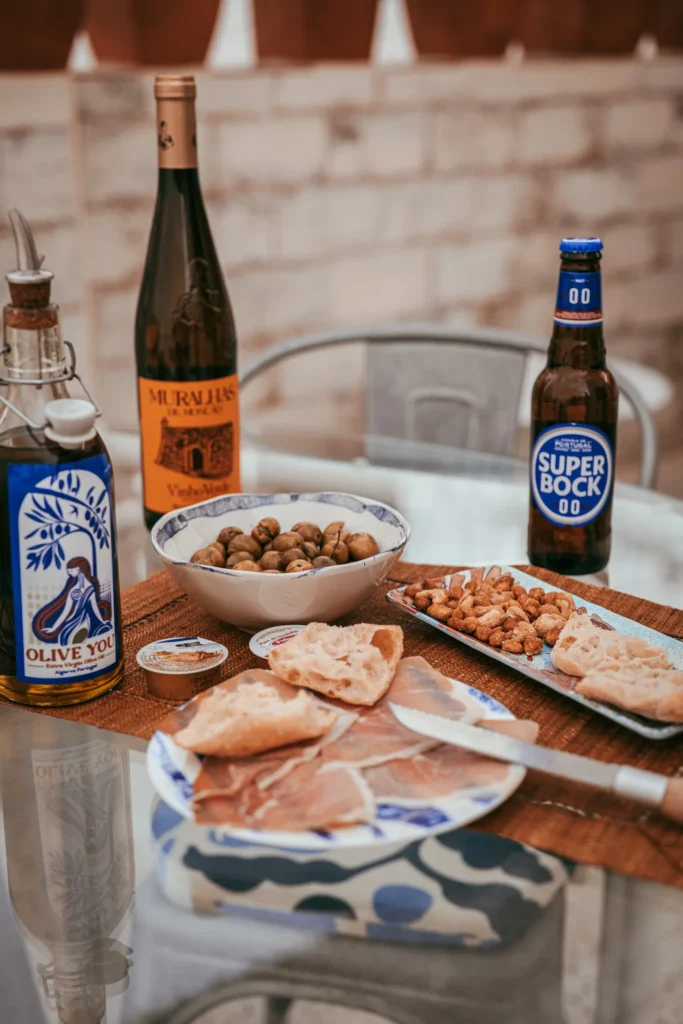 Table set outdoors in Fuseta with bread, ham, olives, roasted almonds, olive oil, a bottle of Murallas de Moncao white wine, and a Super Bock beer, Portuguese aperitif atmosphere.