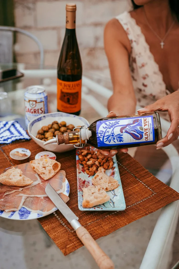 Caroline pours olive oil on a table set with ham, bread, olives, almonds, and drinks during an outdoor meal in Fuseta, Algarve.