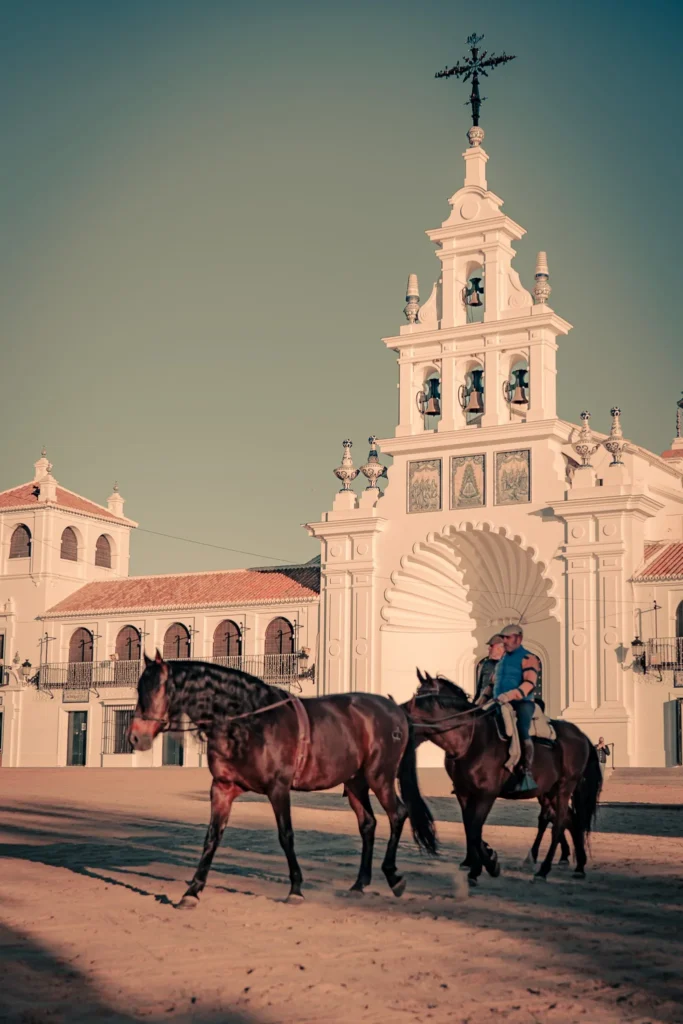 Two horse riders pass in front of the white sanctuary of Virgen del Rocío in El Rocio, under a golden late afternoon light.