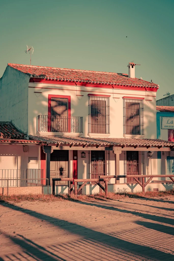 Facade of a traditional Andalusian house in El Rocio, illuminated by the golden evening light, with window grilles and a sandy floor in the foreground.
