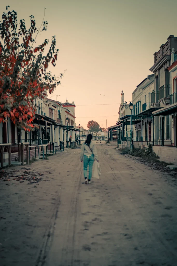 Main street of El Rocio in Andalusia, with a woman walking alone on the sandy path at sunset, surrounded by traditional houses.