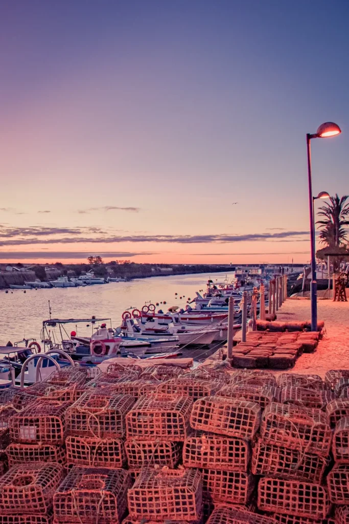 Casiers de pêche empilés sur le quai de Fuseta au coucher de soleil, avec des bateaux amarrés le long de la lagune dans l'Algarve au Portugal.