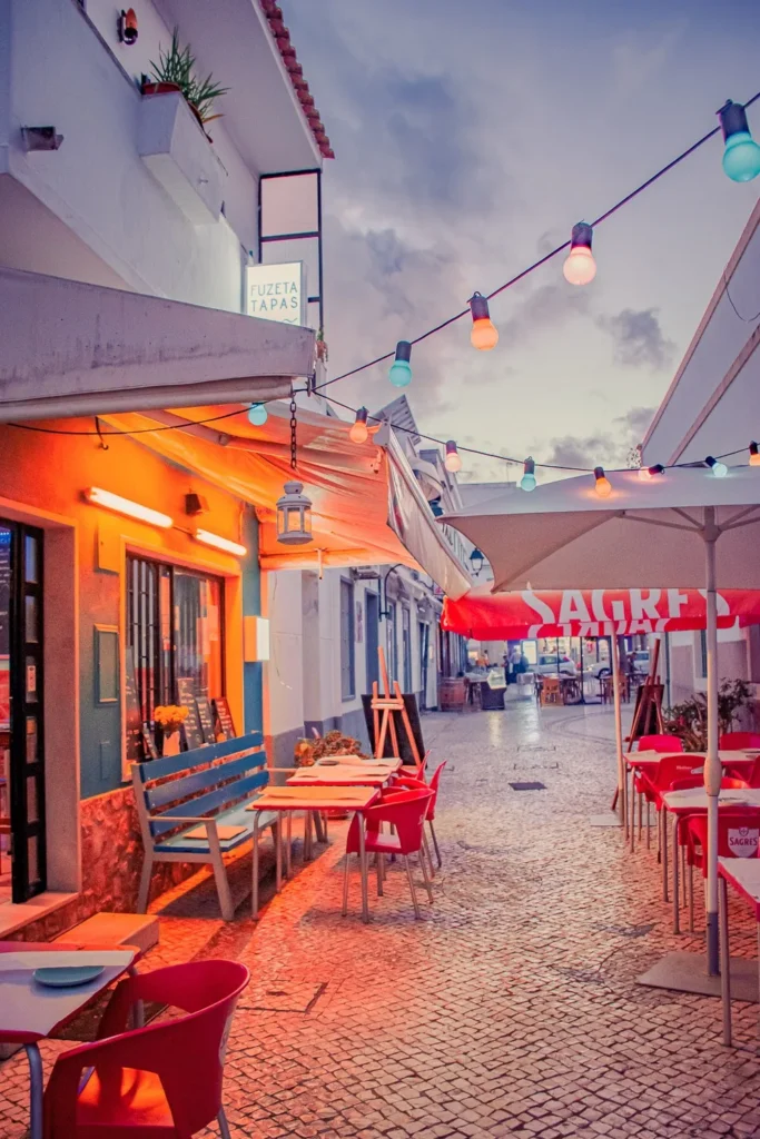 Terrasse de restaurant colorée dans une ruelle pavée de Fuseta, Algarve, au Portugal, en soirée de novembre 2022. Les tables sont vides et des guirlandes lumineuses décorent l'espace.