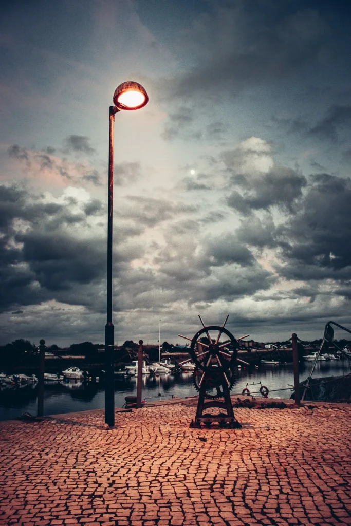 Un quai pavé à Fuseta, Algarve, au Portugal, avec un lampadaire allumé au crépuscule et une grande roue de gouvernail en métal, des bateaux amarrés visibles en arrière-plan sous un ciel nuageux.
