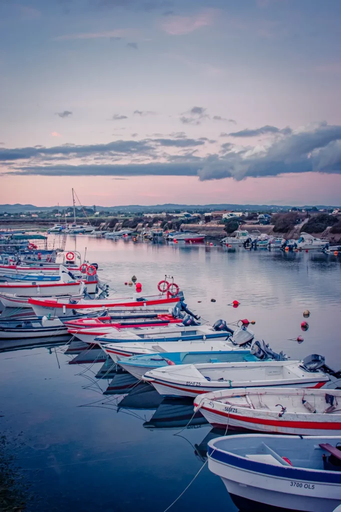 Barques de pêche alignées dans le port de Fuseta en Algarve au coucher du soleil, avec des reflets sur l'eau calme et un ciel nuageux en arrière-plan.