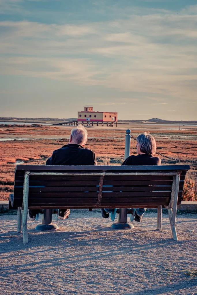 Couple de personne sagées assis sur un banc, contemplant le paysage de lagune à Fuseta dans l'Algarve, avec un bâtiment sur pilotis en arrière-plan sous une lumière de fin de journée.
