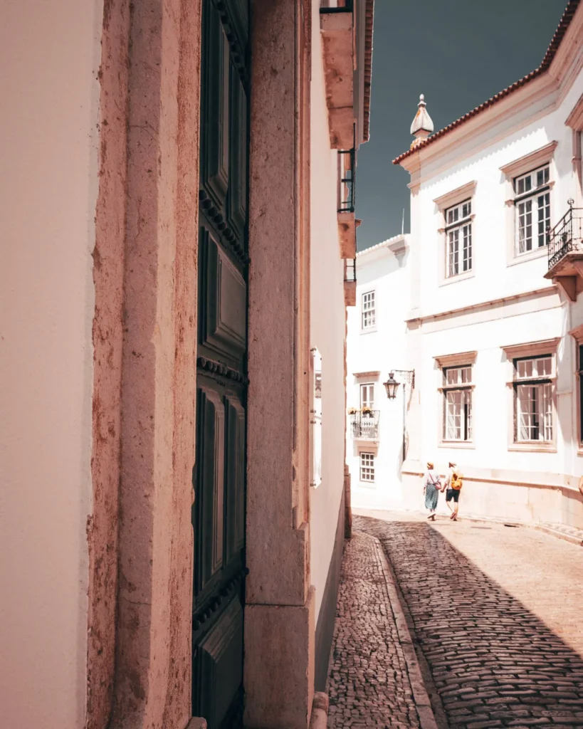 Ruelle pavée bordée de bâtiments blancs traditionnels à Faro, sous une lumière douce de novembre. Deux personnes marchent au loin dans cette rue typique de l'Algarve.