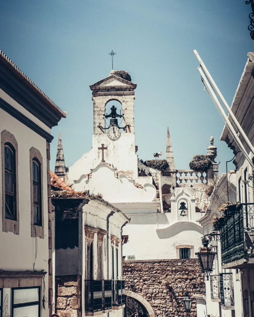 Rue étroite du centre historique de Faro avec vue sur une église blanche et son clocher, sous un ciel bleu en Algarve, Portugal.