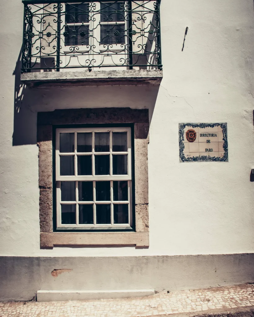 Façade d'un bâtiment historique à Faro avec une fenêtre à petits carreaux, un balcon en fer forgé et une plaque indiquant 'Directoria de Faro'.