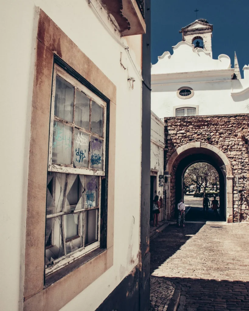 Ruelle pavée de Faro avec une fenêtre taguée au premier plan et une église blanche en arrière-plan, sous un ciel bleu. Quelques personnes sont visibles à l'ombre de l'arche.