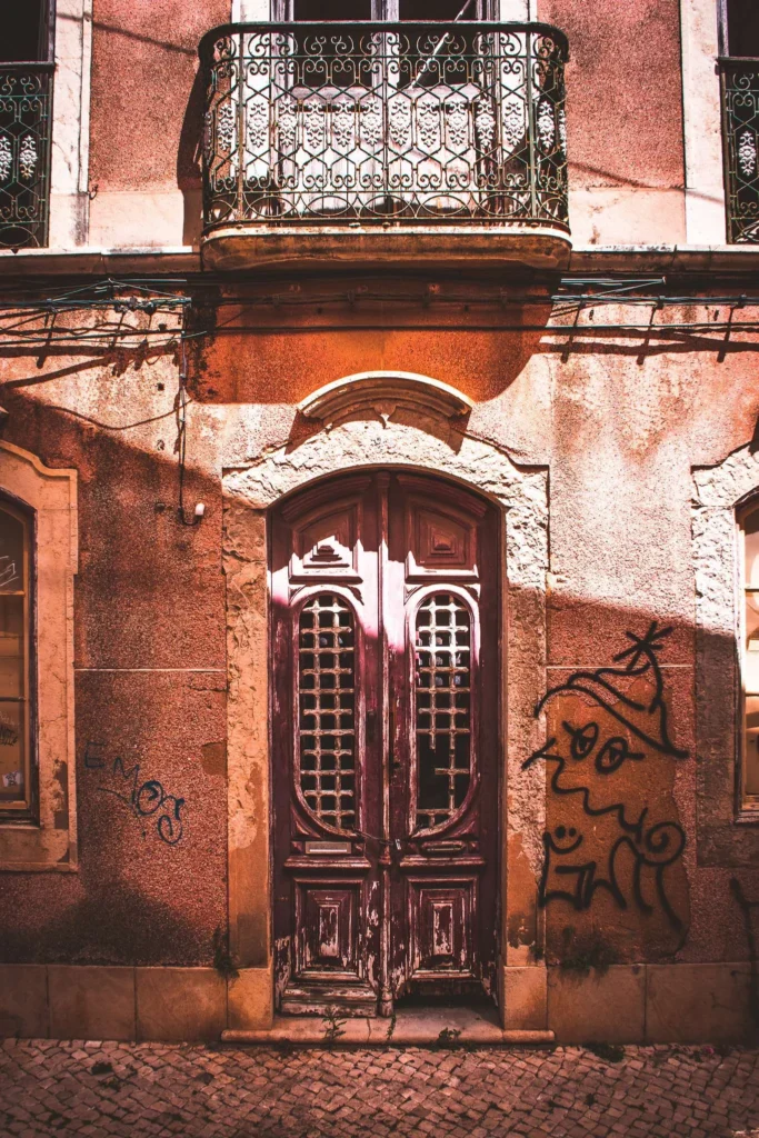 Façade d'un immeuble ancien à Faro, Algarve, avec une porte en bois usée, un balcon en fer forgé et des graffitis sur les murs. La lumière du soleil met en valeur la texture des pierres et du crépi.