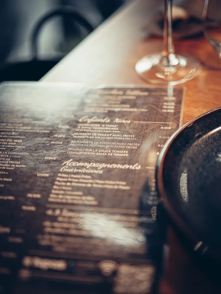 Speisekarte des Restaurants 8 Tapas in Faro auf einem Holztisch, mit einem Weinglas und einem Teller im Vordergrund sichtbar.