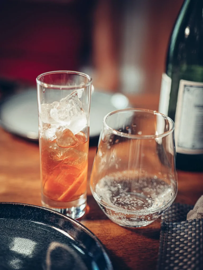 Zwei Gläser auf einem Holztisch in einem Restaurant, eines mit einem Martini Rosso Schweppes mit Eiswürfeln und Zitrusfrüchten, das andere mit Sprudelwasser.