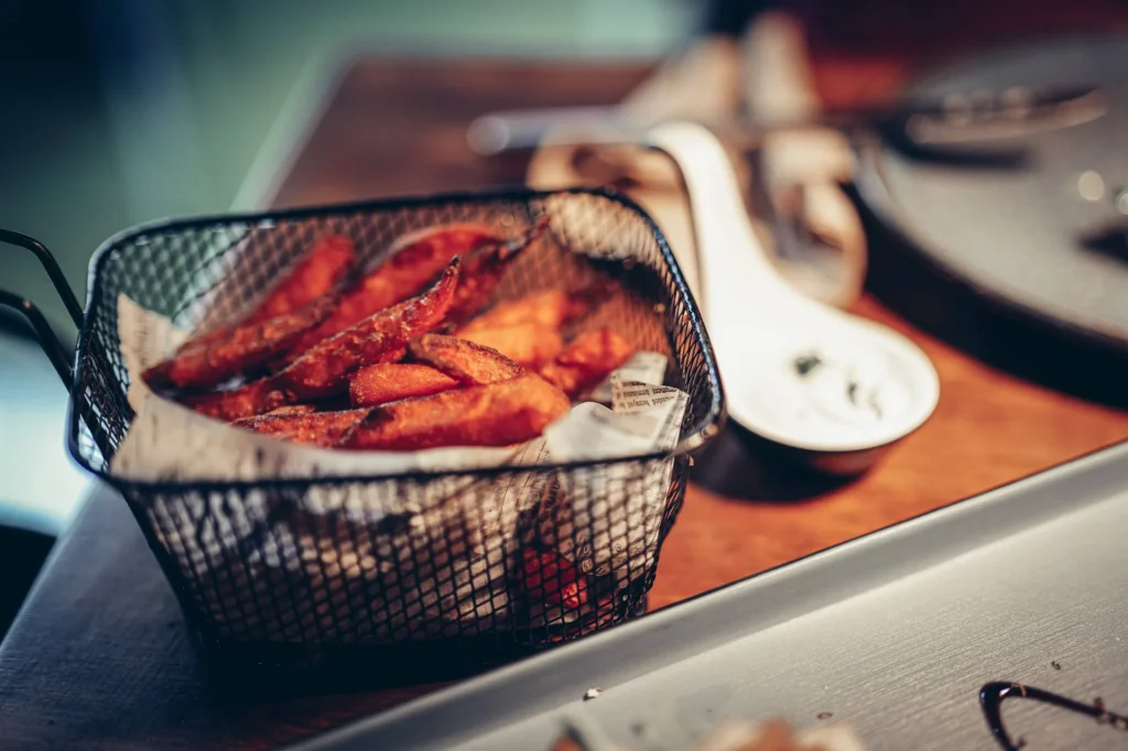 Süßkartoffel-Pommes frites, serviert in einem Metallkorb auf einem Restauranttisch, mit einer Sauce als Beilage. Typische warme Atmosphäre eines Tapas-Restaurants in Faro, Algarve.