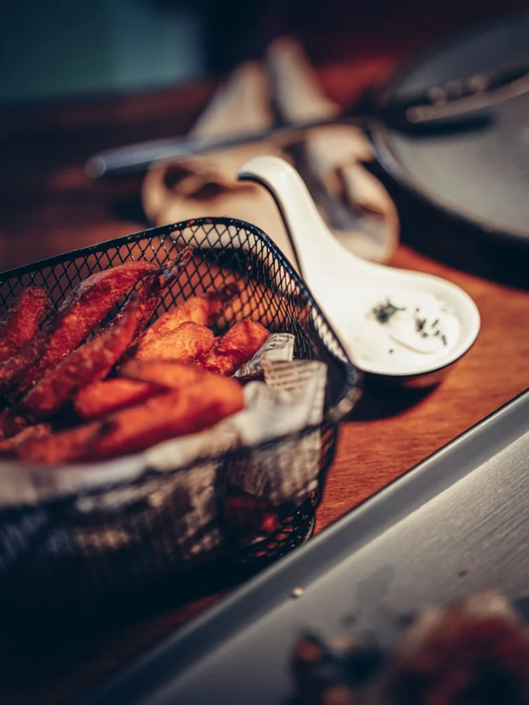Süßkartoffelpommes, serviert in einem Metallkorb auf einem Restauranttisch, mit einer Sauce als Beilage. Gemütliche Atmosphäre, typisch für ein Tapas-Restaurant in Faro, Algarve.