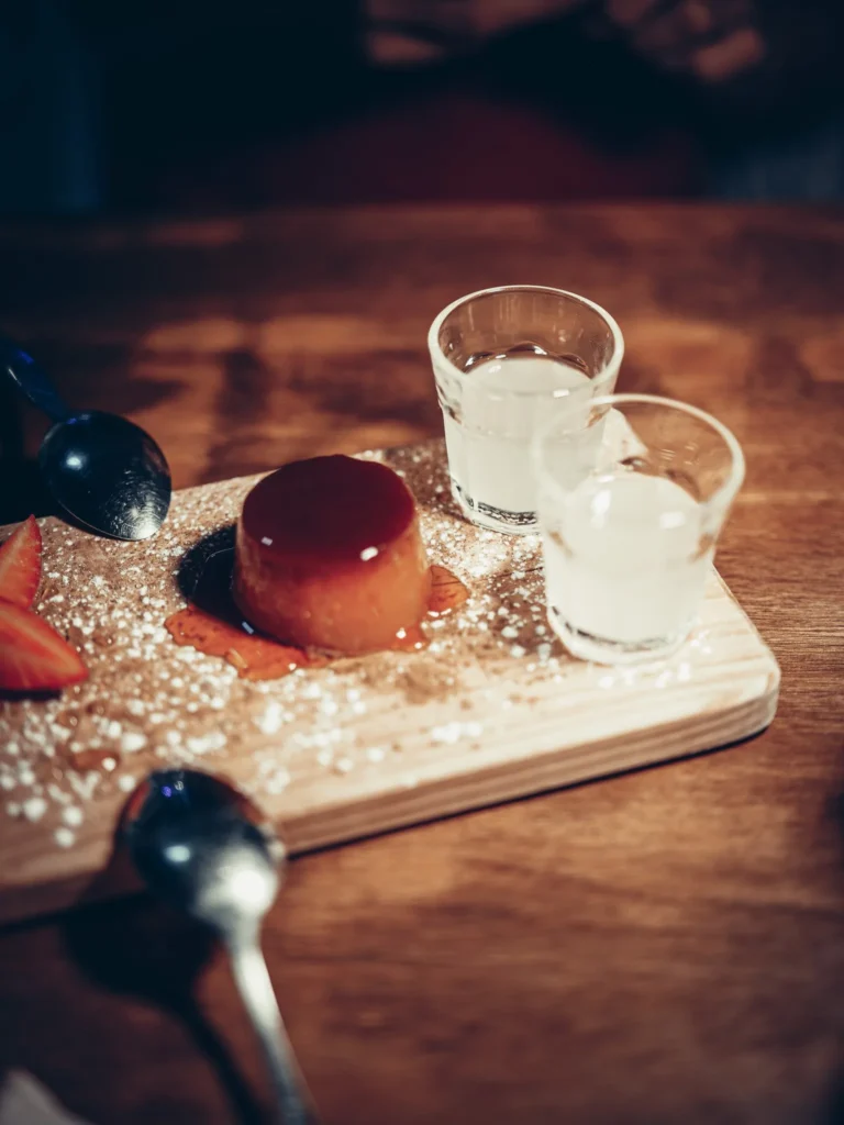 Desserttyp Flan mit Karamell, serviert auf einem Holzbrett mit geschnittenen Erdbeeren, zwei Gläsern Limoncello und Puderzucker, auf einem Tisch im Restaurant 8 Tapas in Faro.
