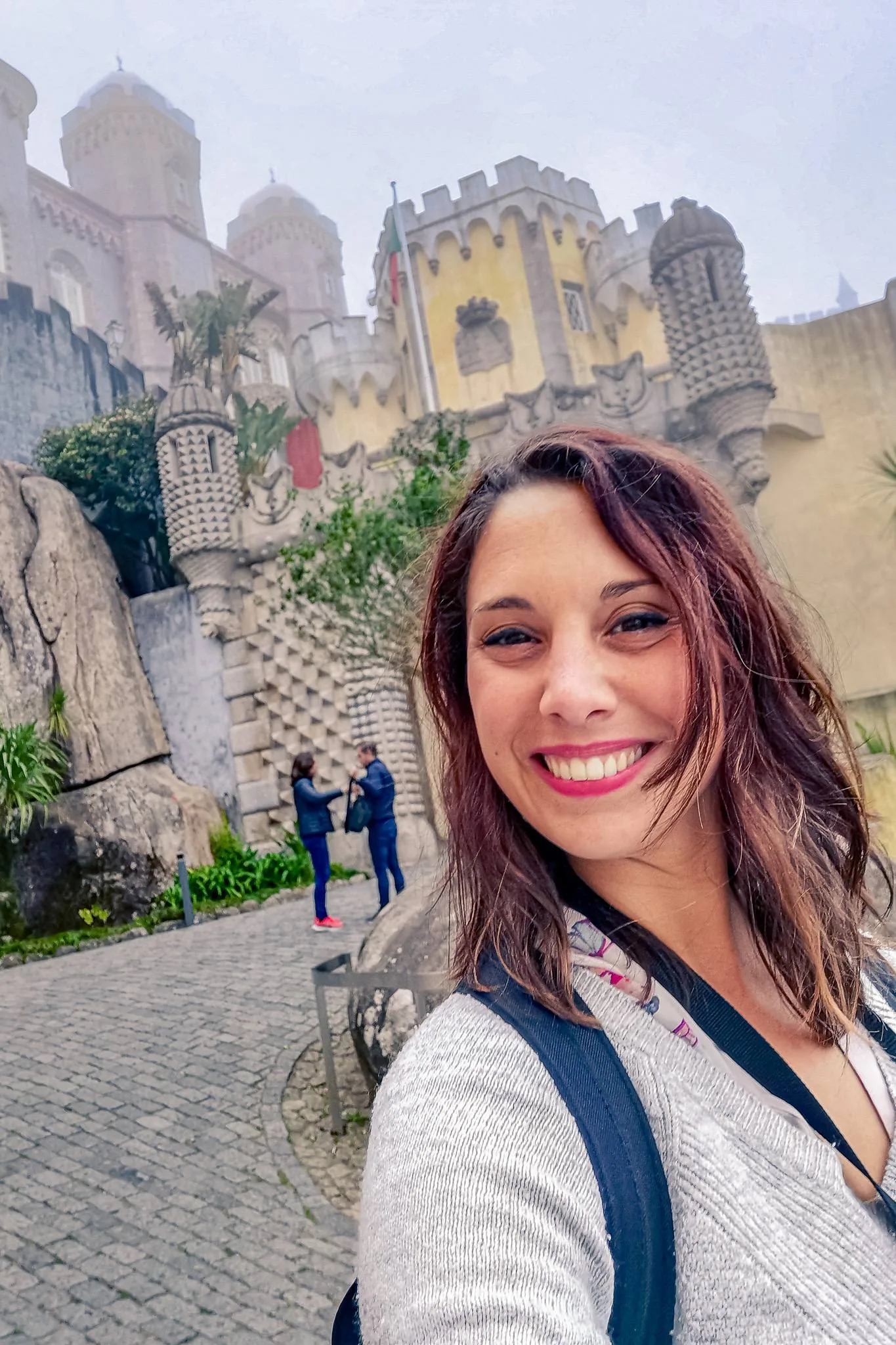 Caroline smiles in front of the colorful and neo-romantic facade of the National Palace of Pena, in a typical misty atmosphere of the Sintra region in February.