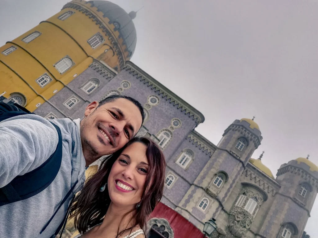 Caroline and Xavier pose in front of the colorful facade of the National Palace of Pena under a gray sky, in Portugal in February 2022.