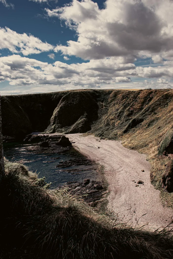 Small pebble beach nestled at the foot of high green cliffs at Dunnottar