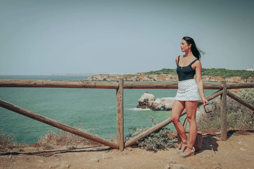 Caroline standing by a wooden barrier on a cliff overlooking the Atlantic Ocean in Portimao, under the May sun.