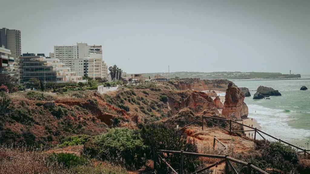 Ochre cliffs overlooking the beach and the Atlantic Ocean in Portimao, with modern buildings in the background under a clear sky.