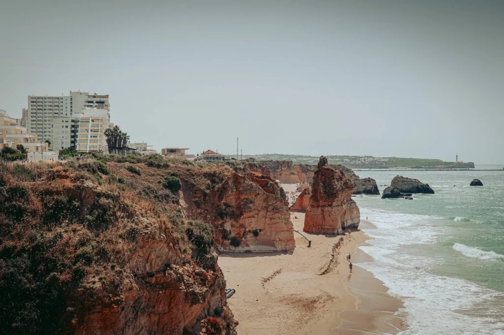 Ochre cliffs and iconic rock formations line a beach in the Algarve at Portimao, with modern buildings in the background and the Atlantic Ocean under a slightly overcast sky.