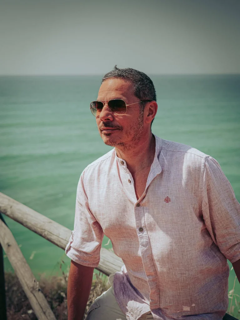 Xavier in a light shirt and sunglasses in front of the Atlantic Ocean, on a coastal path in Portimao in the Algarve, Portugal, under summer light.