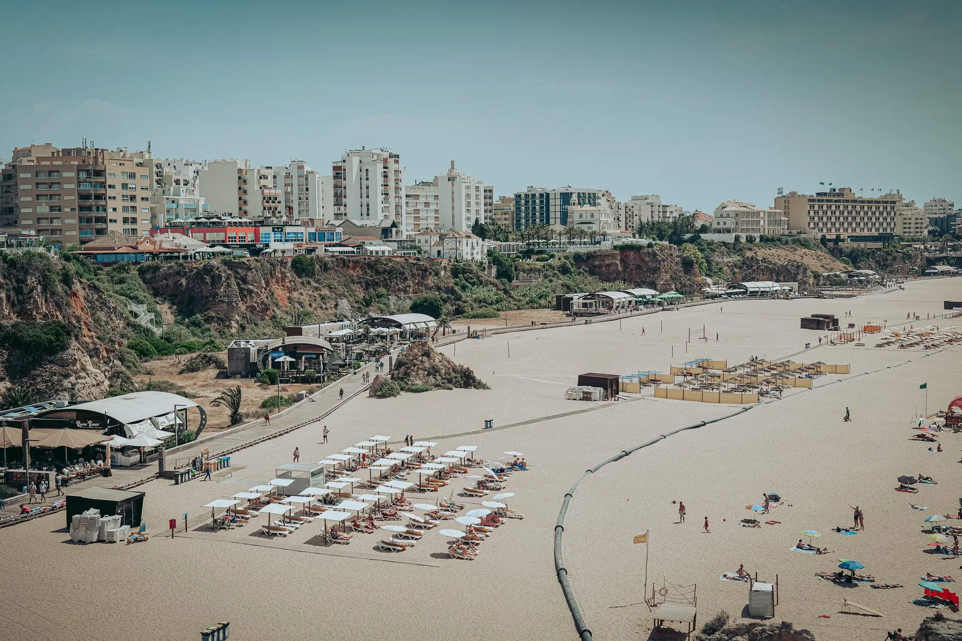 Large sandy beach in Portimao with aligned umbrellas, a few holidaymakers, and buildings in the background, under a clear sky in the Algarve at the end of May.