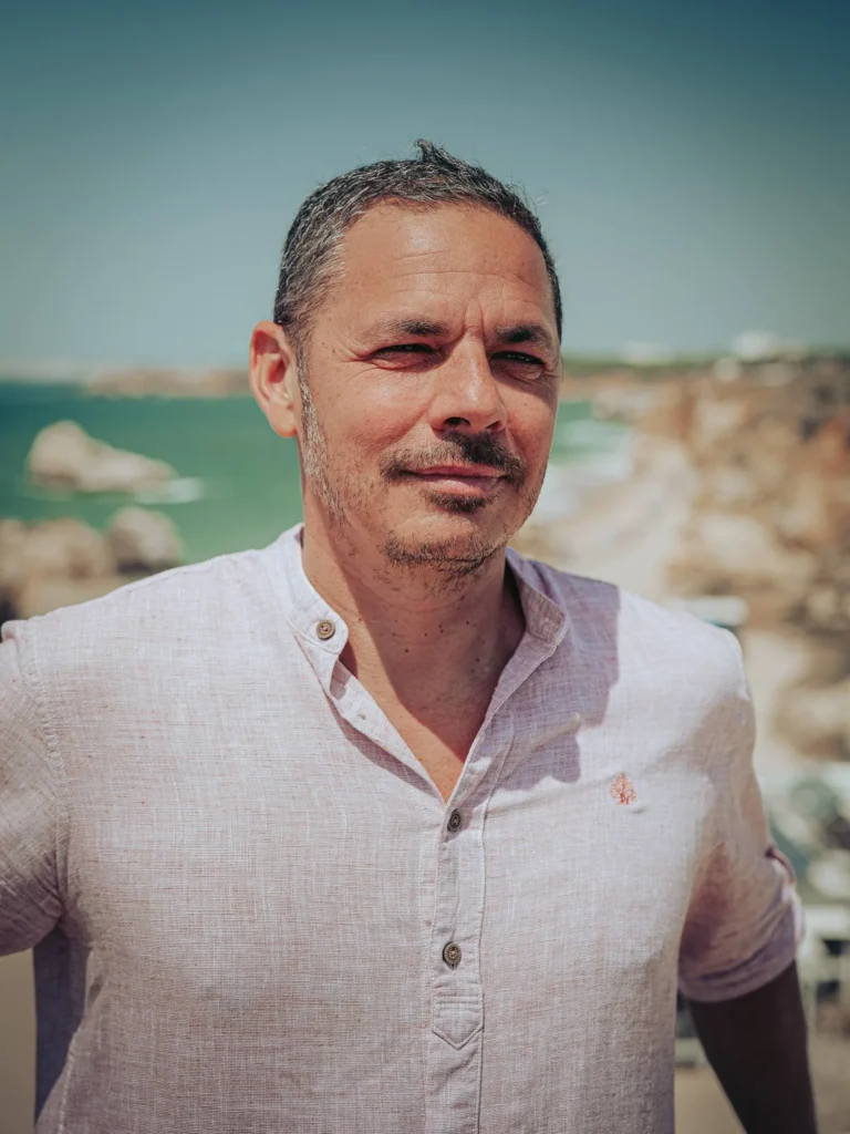 Xavier in a light shirt in front of the cliffs and the ocean in Portimao, Algarve, under a sunny sky in May 2026.