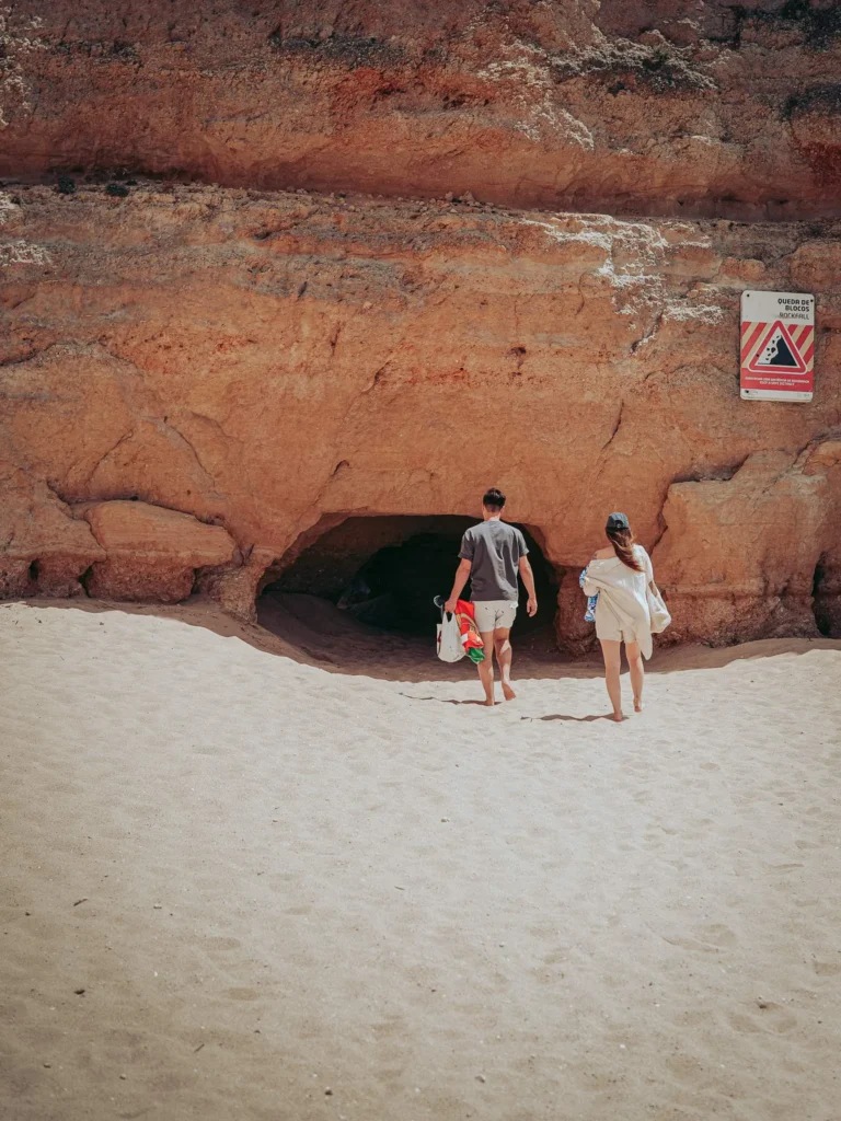 Caroline and Xavier enter a small cave carved into a sandy cliff on a beach in the Algarve, beneath a warning sign.