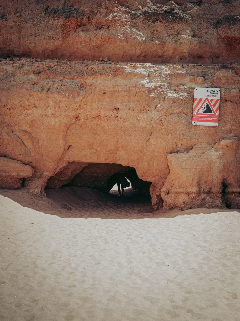 Cave carved into a ochre rock cliff on a beach in the Algarve, with a warning sign against falling rocks.