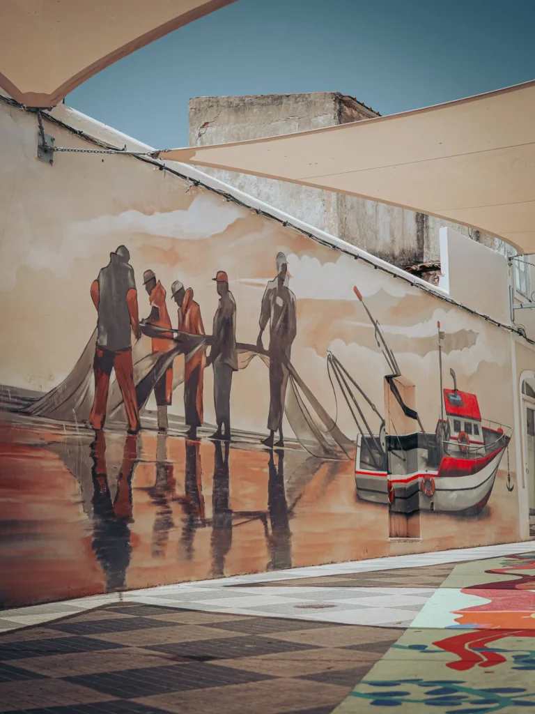 Mural depicting fishermen pulling a net and a fishing boat, on an exterior wall in Portimao under a clear sky.