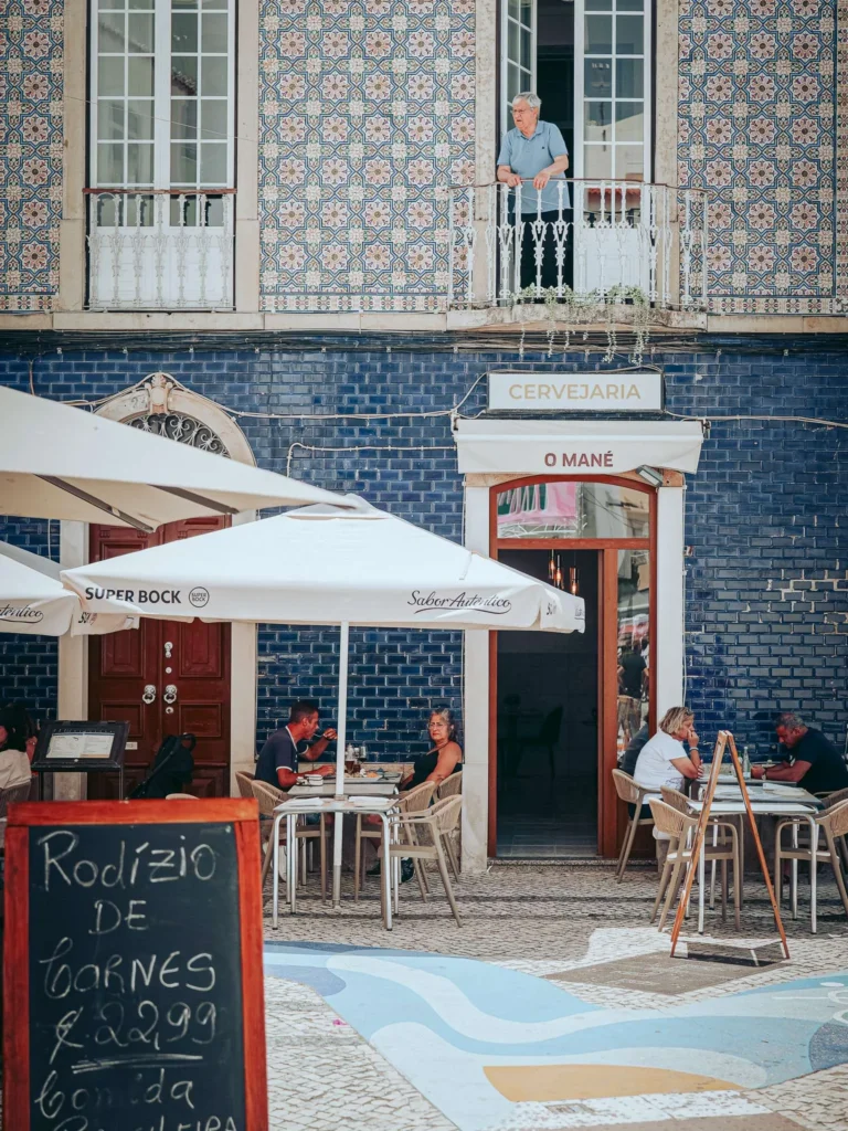 Restaurant terrace in Portimao with customers seated under parasols, in front of a facade decorated with typical blue azulejos from the Algarve, a man stands on the balcony above.