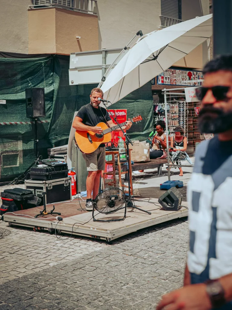 Xavier plays guitar and sings on a small street stage in Portimao, under a large white umbrella, on a sunny day.