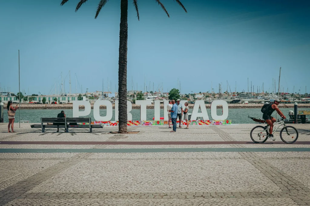 View of the Portimão harbor with the large white letters of the city's name along the quay, pedestrians and a cyclist in the foreground, under a clear sky.