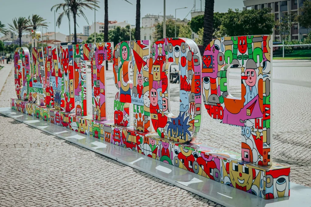 Colorful and artistic lettering 'Portimao' installed on a cobblestone promenade, with palm trees and buildings in the background under the Algarve sun.