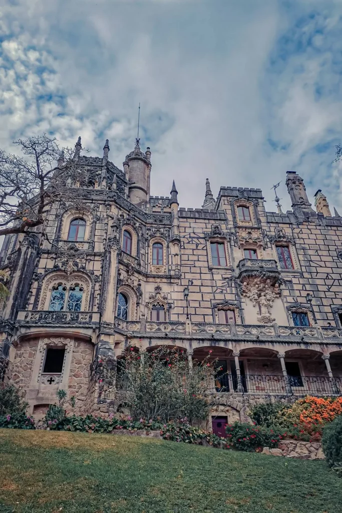Facade of the Quinta da Regaleira palace in Sintra, surrounded by blooming gardens under a cloudy sky in March 2022.