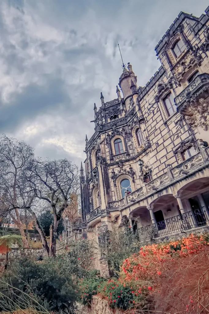 Facade of the Quinta da Regaleira palace in Sintra, surrounded by blooming gardens under a cloudy sky in March 2022.