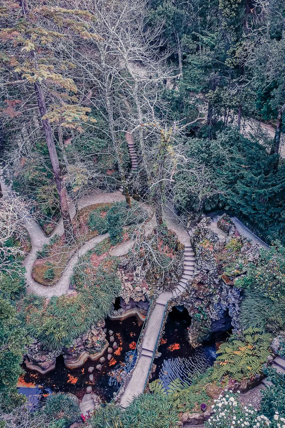 Aerial view of the labyrinthine gardens of Quinta da Regaleira in Sintra, featuring winding paths, stone staircases, and a pond surrounded by vegetation in March.