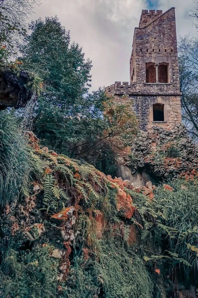 Stone tower covered in vegetation at Quinta da Regaleira, Sintra, photographed in March 2022. The atmosphere is mysterious and natural, with an abundance of ferns and moss.